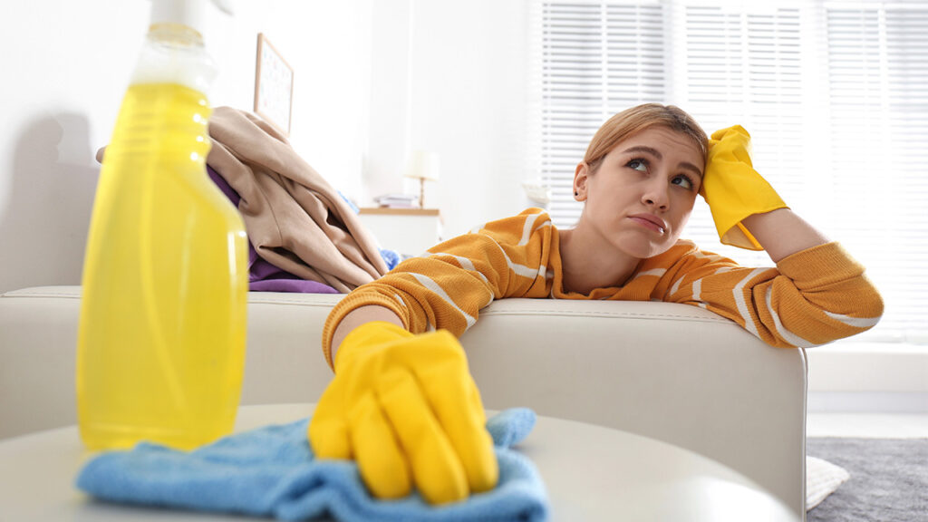 A woman wearing yellow cleaning gloves looks tired and frustrated while surrounded by cleaning supplies in a bright room.