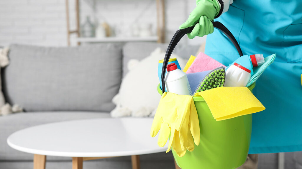 A person in green gloves holds a bucket filled with cleaning supplies, including spray bottles and cloths, in a modern
